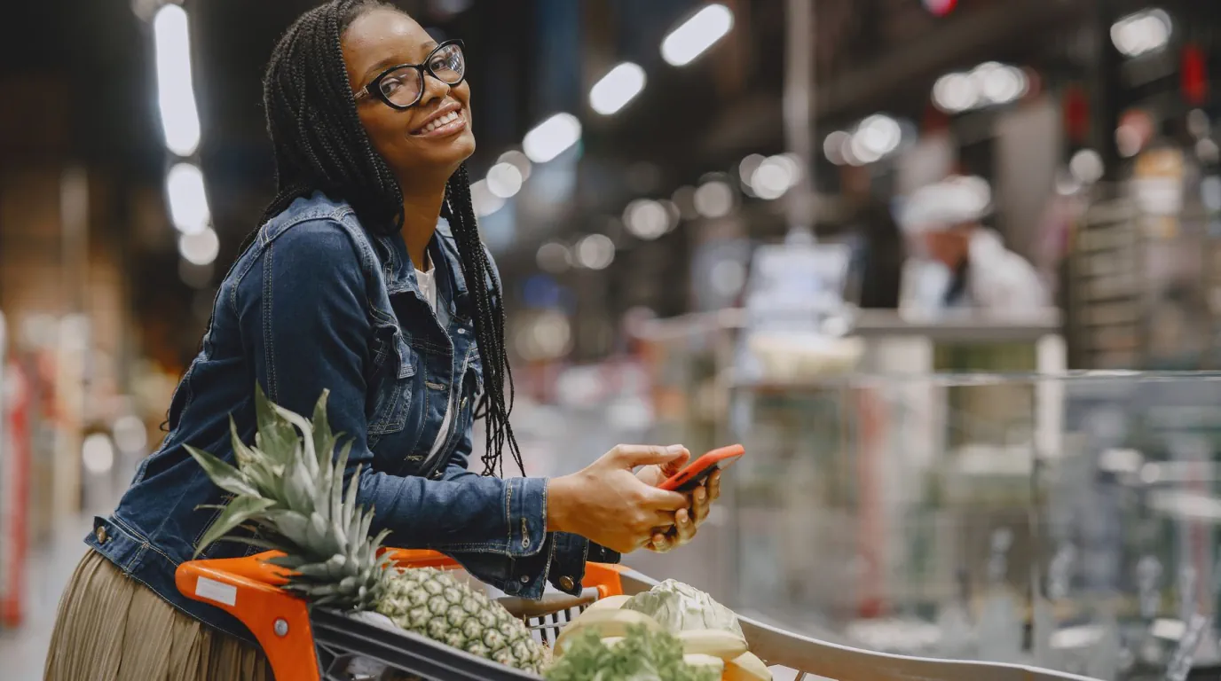 A shopper checks her phone beside a cart filled with fresh groceries.